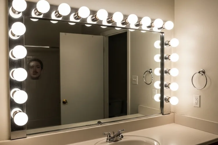 Outdated Hollywood globe vanity lights around bathroom mirror, bright exposed round bulbs causing glare, harsh reflections on face, builder grade bathroom