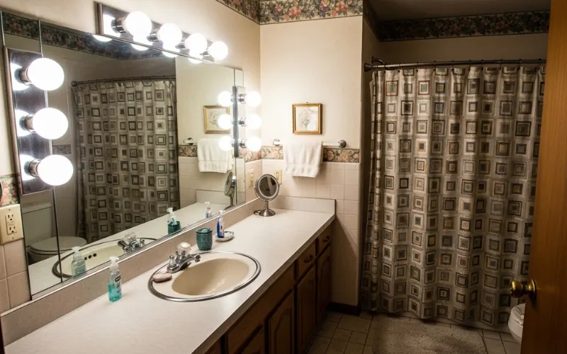 Outdated 90s bathroom with Hollywood globe vanity lights around mirror, harsh glare, uneven lighting shadows, builder grade style.