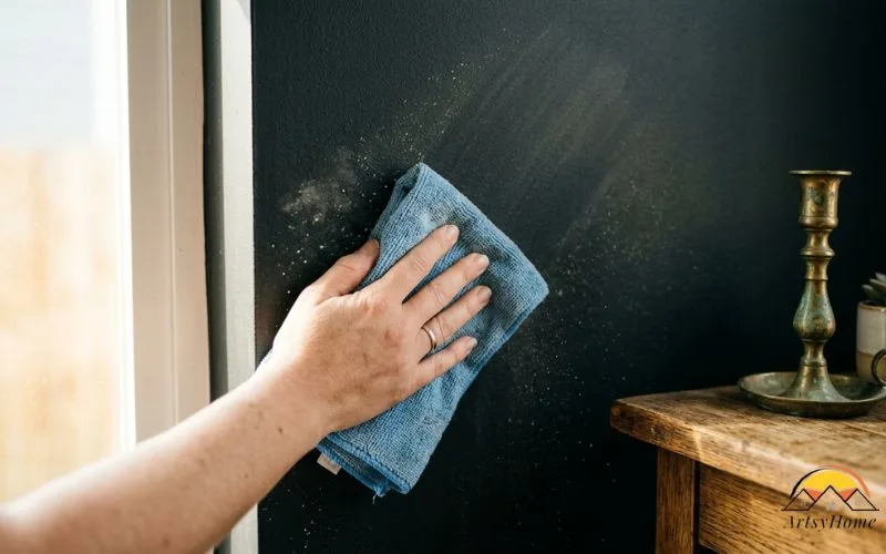 Realistic close-up photograph of a hand holding a microfiber cloth dusting a deep matte green wall, catching visible dust in raking side light to illustrate the maintenance point