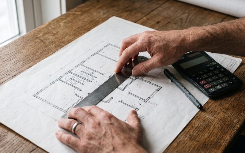 Photorealistic close-up of an architect or contractor measuring a real floor plan on a wooden desk.