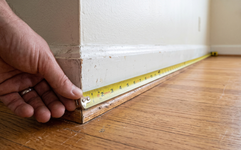 Ultra-realistic close-up photograph of a steel tape measure lying flat directly along the floor from one wall to the opposite wall